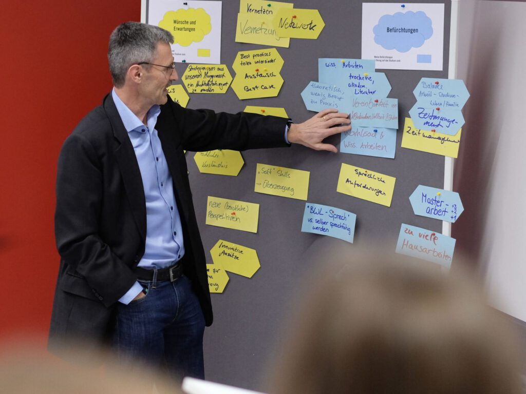 Prof. Stefan Schaltegger, der Studiengangsleiter des MBA Sustainability Management, steht vor einer Pinnwand. Darauf sind gelbe Karten mit den Erwartungen und blaue Karten mit den Befürchtungen der Studierenden zu erkennen.