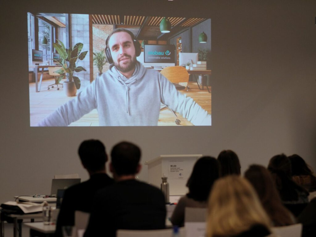 Pascal Schwarz von elobau hält einen Vortrag bei der Einführungspräsenz des MBA Sustainability Management. Da der Vortrag digital stattfand, sitzen die Studierenden im Raum und schauen auf ein auf die Wand projiziertes Video.