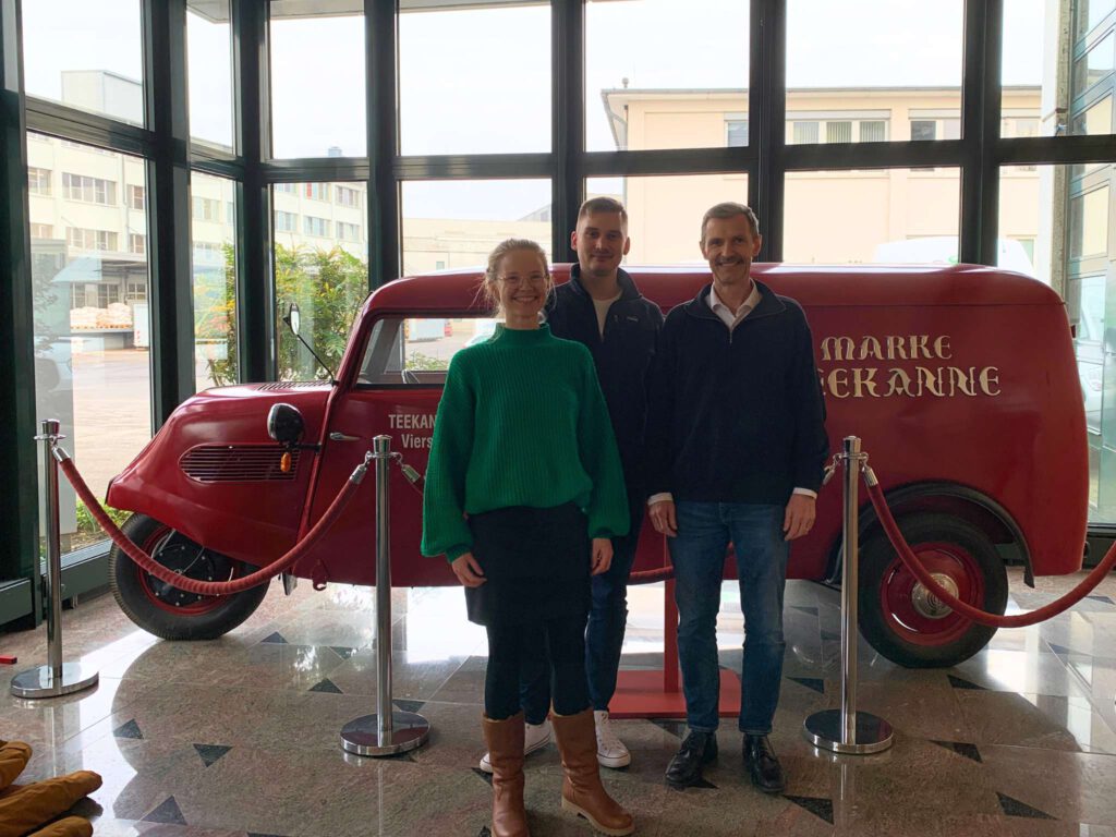 Prof. Stefan Schaltegger, der Studiengangsleiter des MBA Sustainability Management, und Katharina Guhl und Benjamin Sachs aus dem Team des Centre for Sustainability Management, stehen vor einem roten historischen Auto im Firmengebäude der Teekanne GmbH.