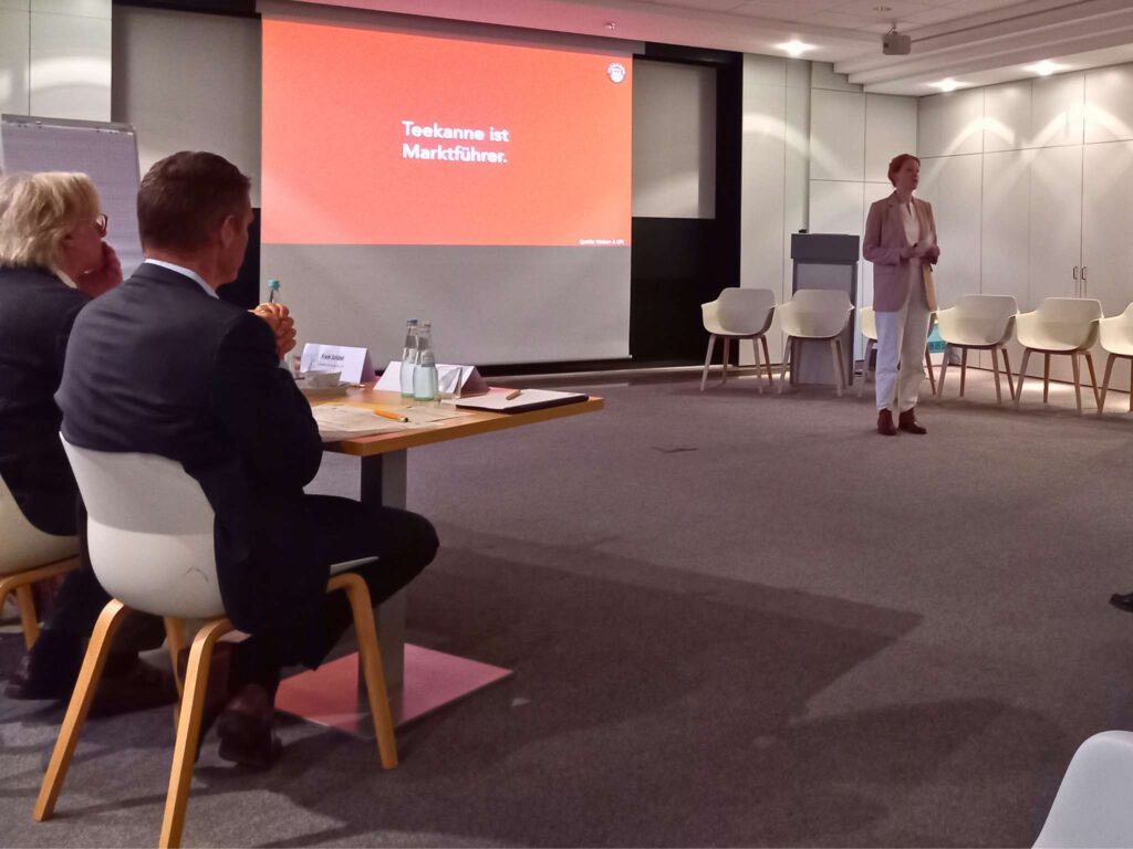 Die HR-Referentin von Teekanne, Annika Schur, hält einen Vortrag. Auf die Leinwand wird eine Präsentation projiziert, auf der "Teekanne ist Marktführer" auf orangenem Hintergrund steht.