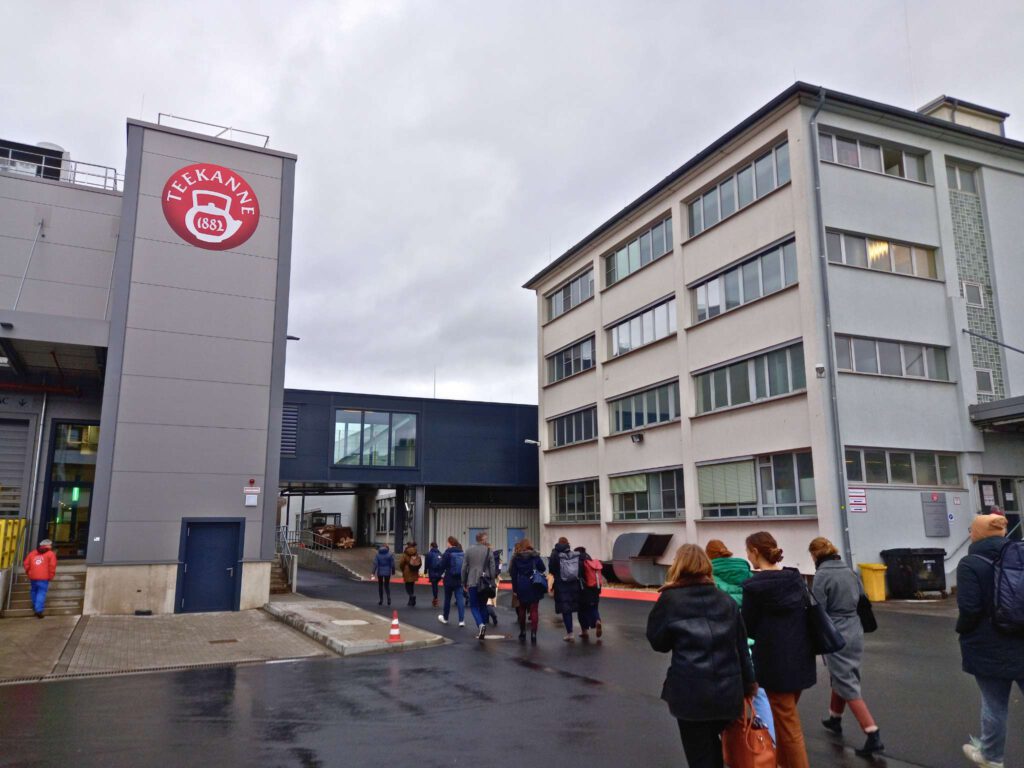 Die Studierenden des MBA Sustainability Management betreten das Firmengelände der Teekanne Holding GmbH & Co. KG. An einem Gebäude auf der linken Seite ist groß das rote Teekanne-Logo zu erkennen. Der Himmel ist grau.