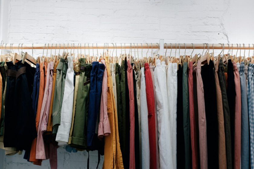 verschiedene bunte Kleidungsstücke hängen auf Kleiderbügeln an einer Holzstange vor einer weißen Wand